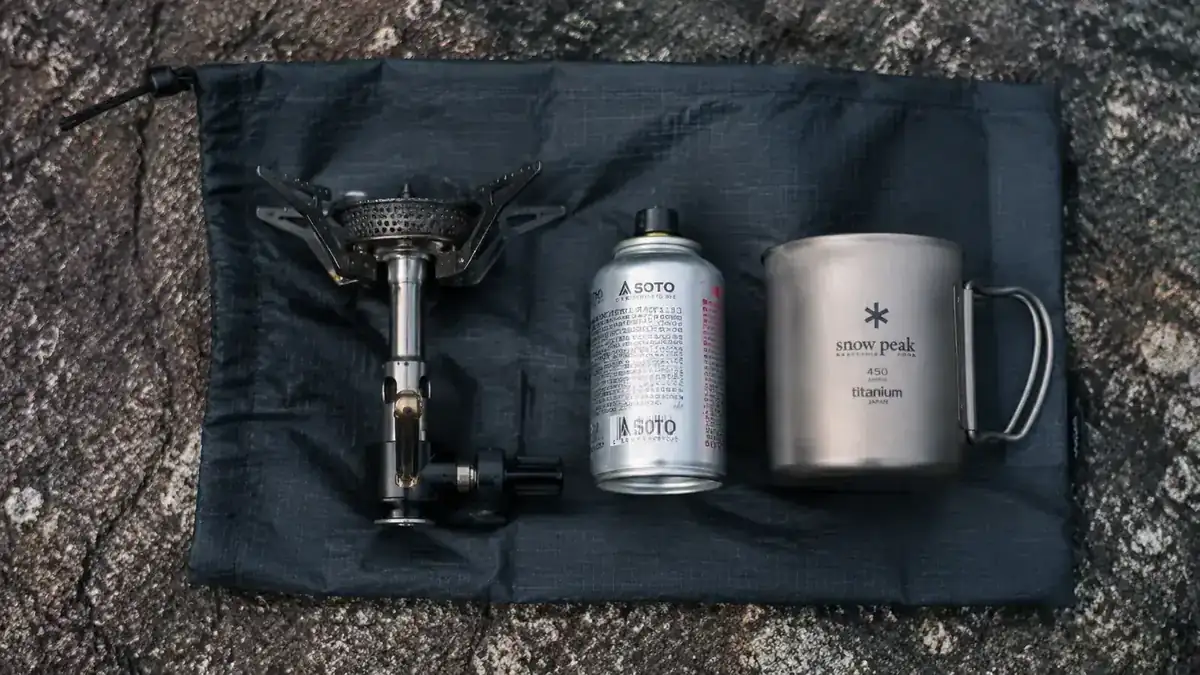 Backpacker's open stuff sack showing minimal cook kit: stove, canister, and titanium mug as the entire cooking system