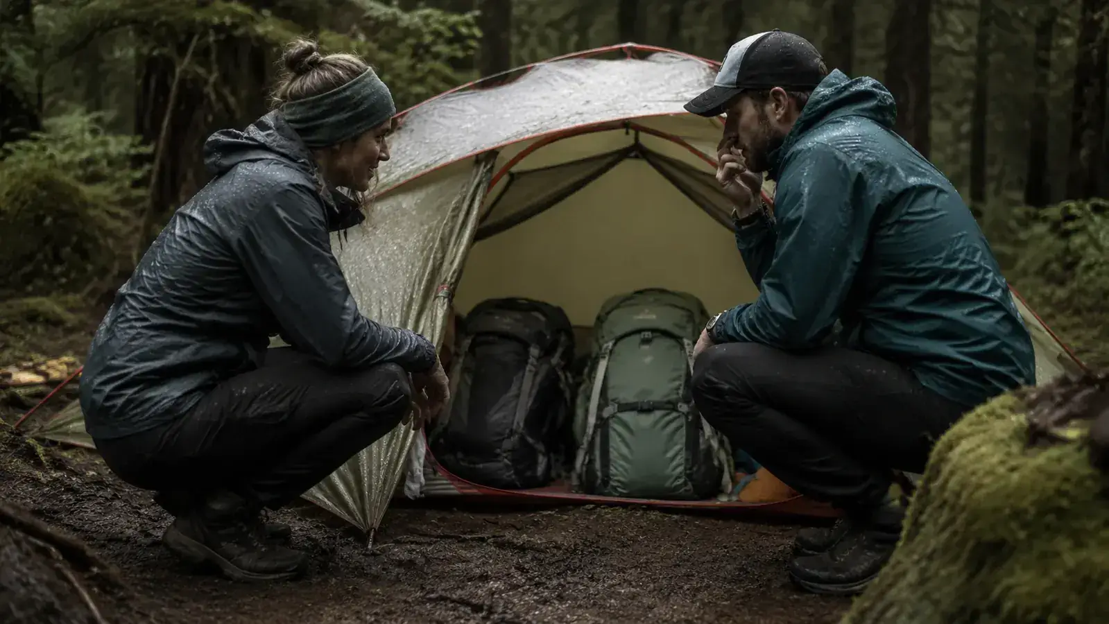 Two campers with full packs trying to fit gear under a small backpacking tent vestibule in the rain