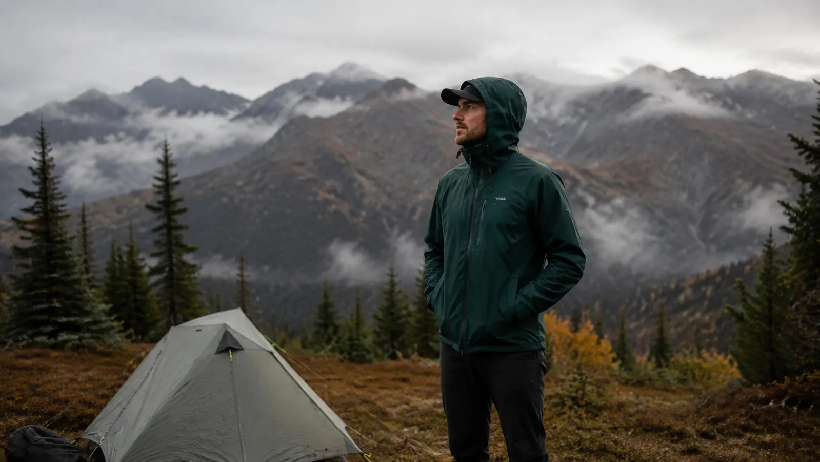 Backpacker in rain gear standing beside a pitched tent at a Cascades trailhead camp, mountains in the distance under low clouds