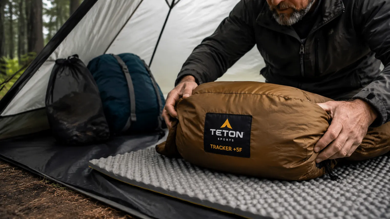 Hand unrolling the TETON Sports Tracker sleeping bag onto a foam sleeping pad inside a tent
