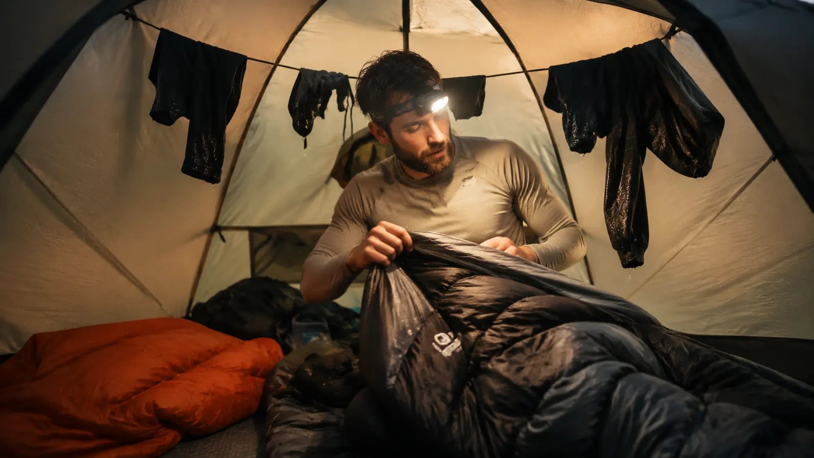 Camper in dry base layer clothes crawling into a mummy sleeping bag inside a tent surrounded by rain gear hung on a cord