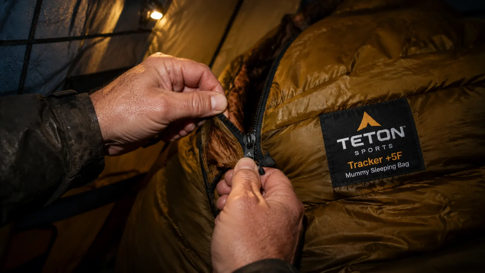 Close-up of a man's hands struggling with a sleeping bag zipper draft tube inside a dark tent, headlamp illuminating the scene