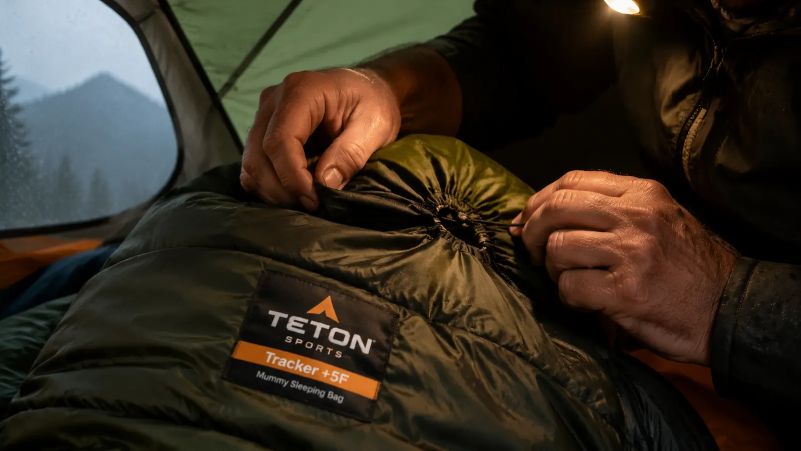 Hands cinching the mummy hood drawcord on the TETON Tracker sleeping bag inside a backpacking tent, headlamp clipped to tent peak above
