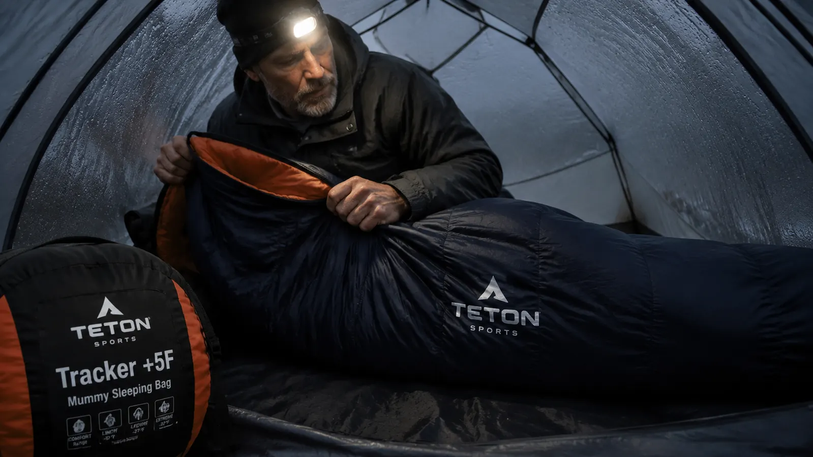 Camper sliding into a green mummy sleeping bag inside a tent with condensation on the interior walls